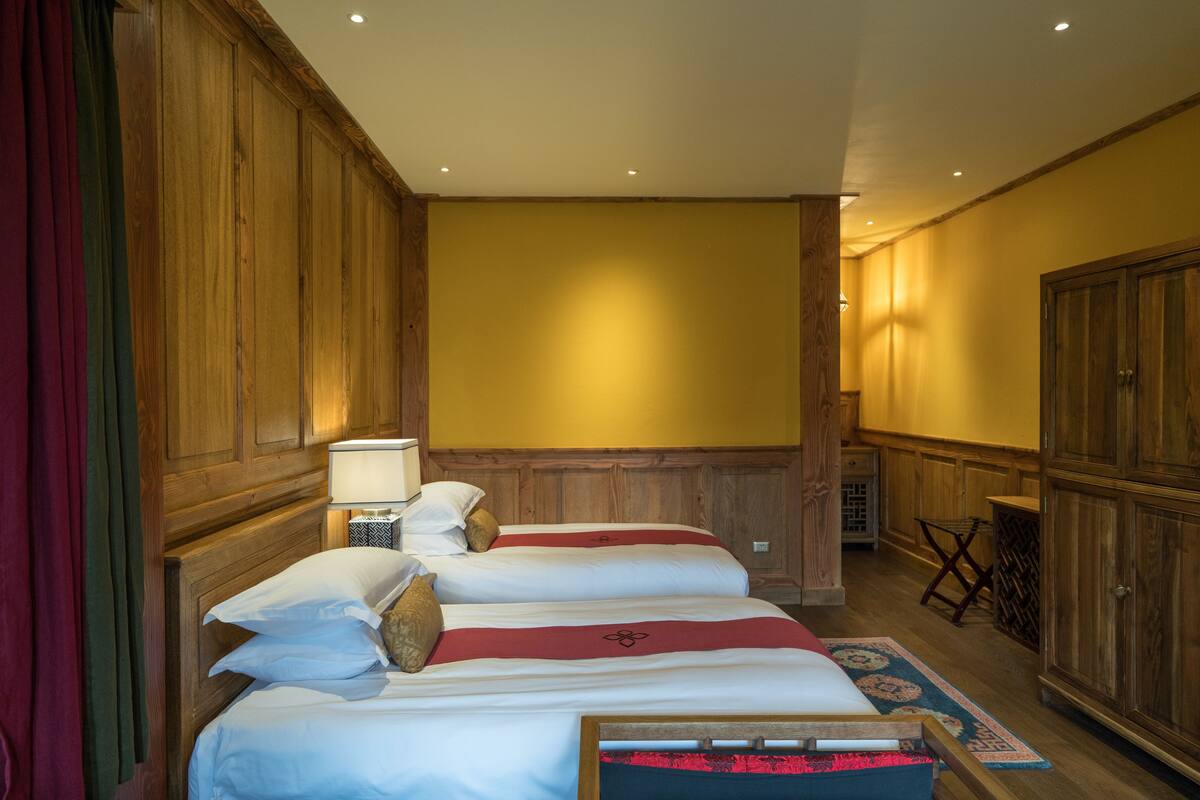 A hotel room with two beds, wooden paneling, a small table with a lamp, and a wardrobe.