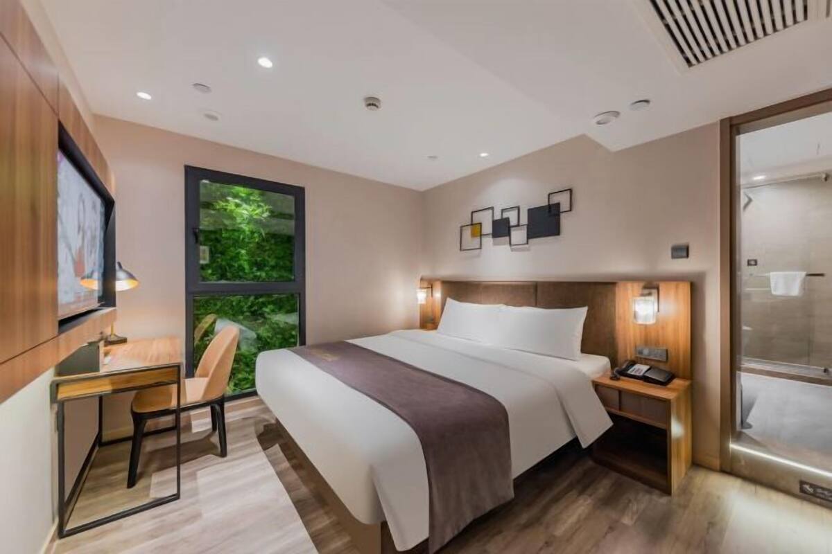 A modern hotel room with a bed, desk, chair, and a large window with a view of greenery.