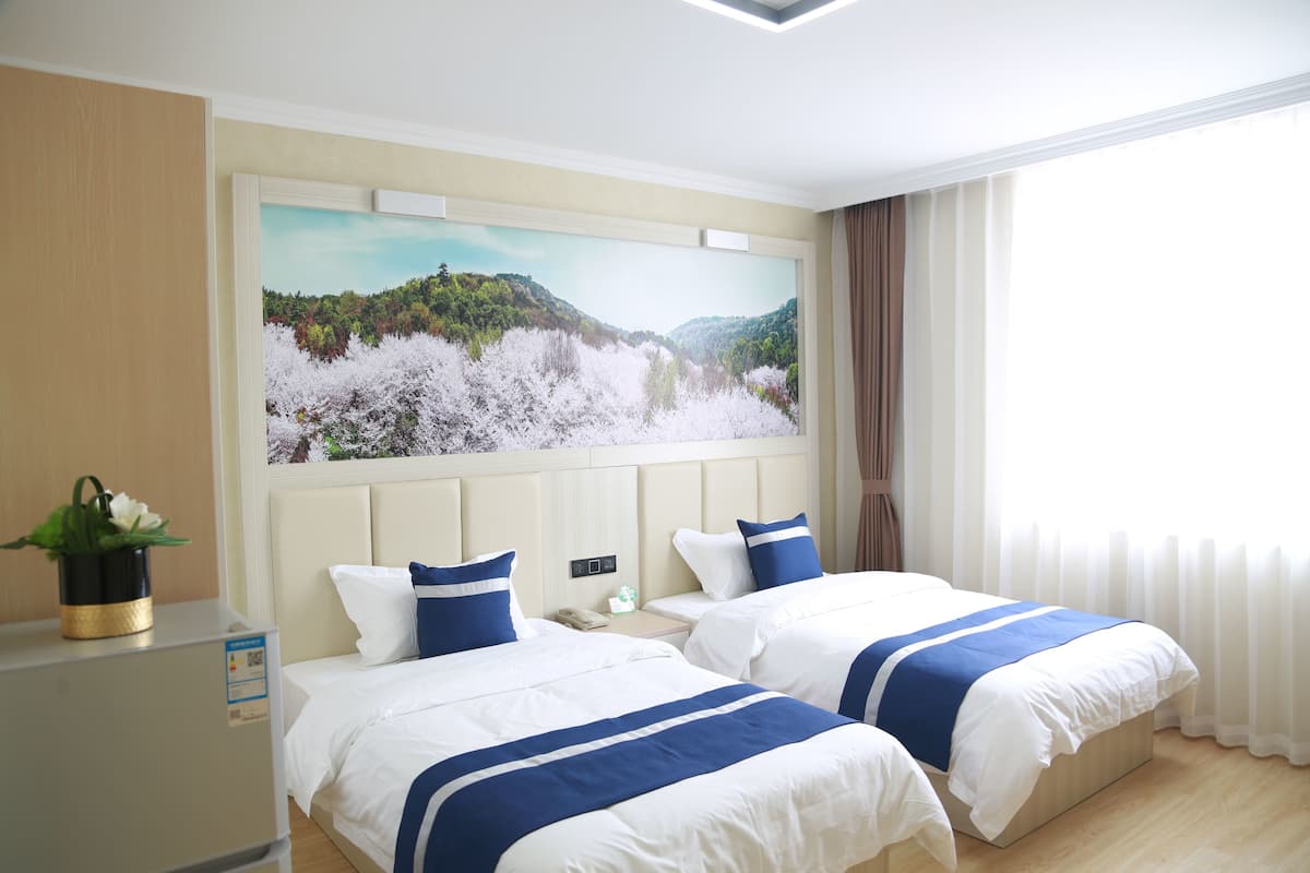 A hotel room with two beds, a large landscape painting on the wall, a bedside table with a phone, and a window with curtains.