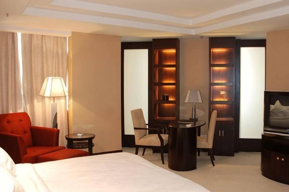 Ein Hotelzimmer mit einem großen Bett, einem roten Sessel, einem Schreibtisch mit Lampe, einem Fernseher und einem Bücherregal mit beleuchteten Ablagen.