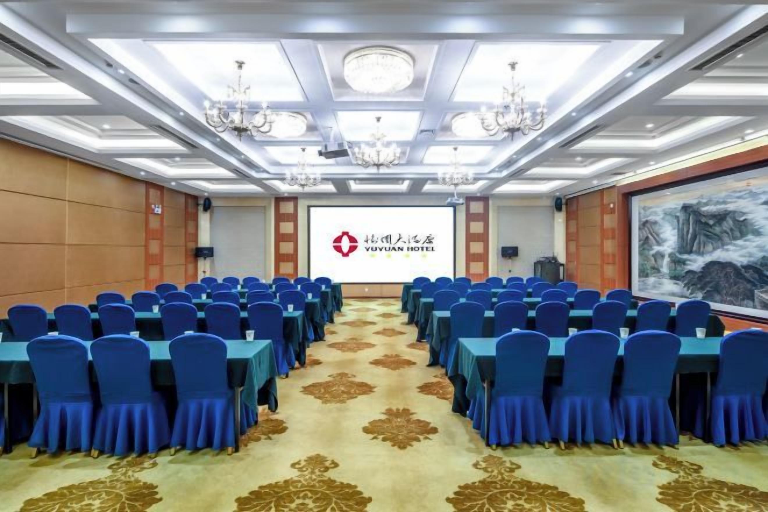 A conference room with rows of blue chairs, a carpet with a floral pattern, chandeliers, and a screen displaying the Yuyuan Hotel logo.
