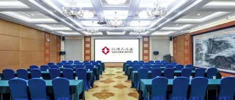Uma sala de conferências com fileiras de cadeiras azuis, um tapete com padrão floral, lustres e uma tela exibindo o logotipo do Hotel Yuyuan.
