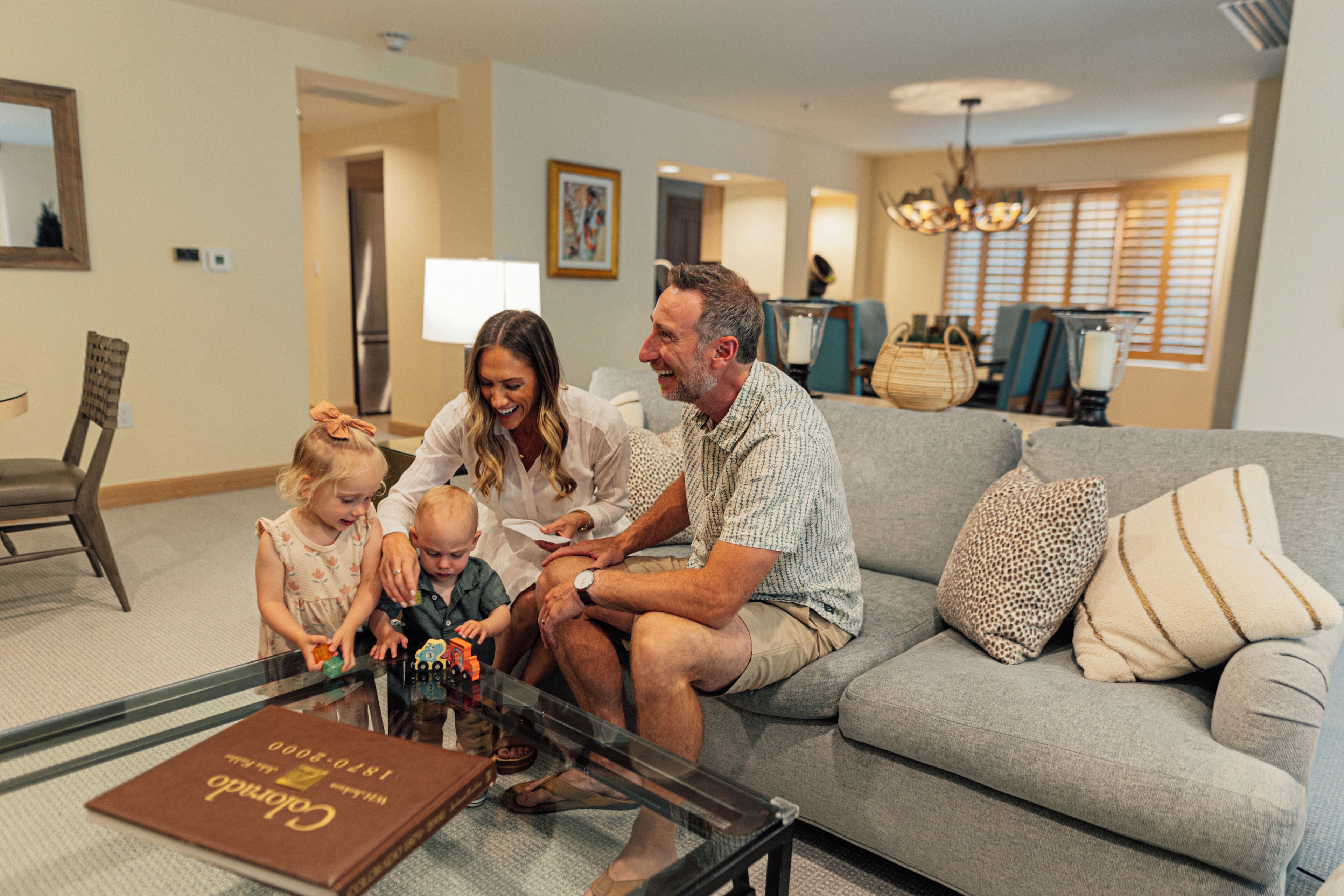 Une famille est assise sur un canapé gris dans un salon bien éclairé, avec une table basse en verre, un coin repas à l’arrière-plan et un lustre au plafond.