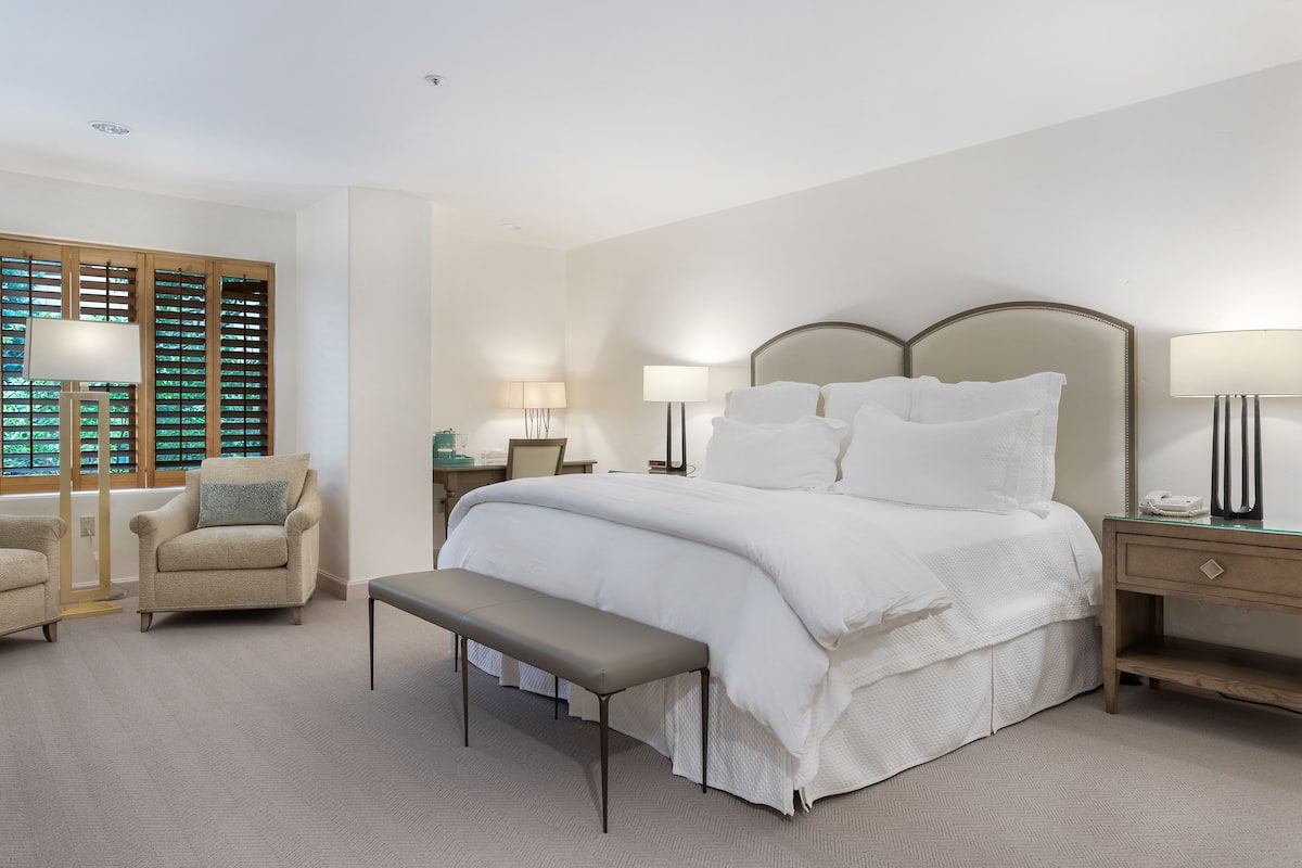A bedroom with a large bed, a chair, a bench, and a nightstand with a lamp.
