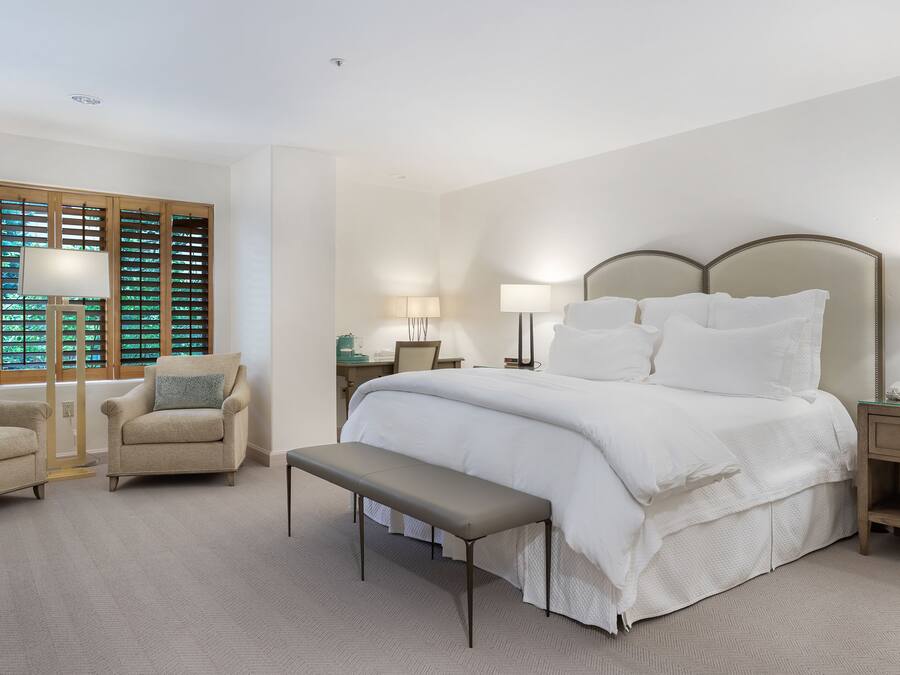 A bedroom with a large bed, a chair, a bench, and a nightstand with a lamp.