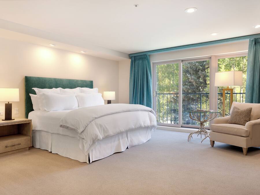 Habitación de hotel con una cama grande, dos mesitas de noche con lámparas, una silla, una pequeña mesa y vistas a árboles a través de la ventana.