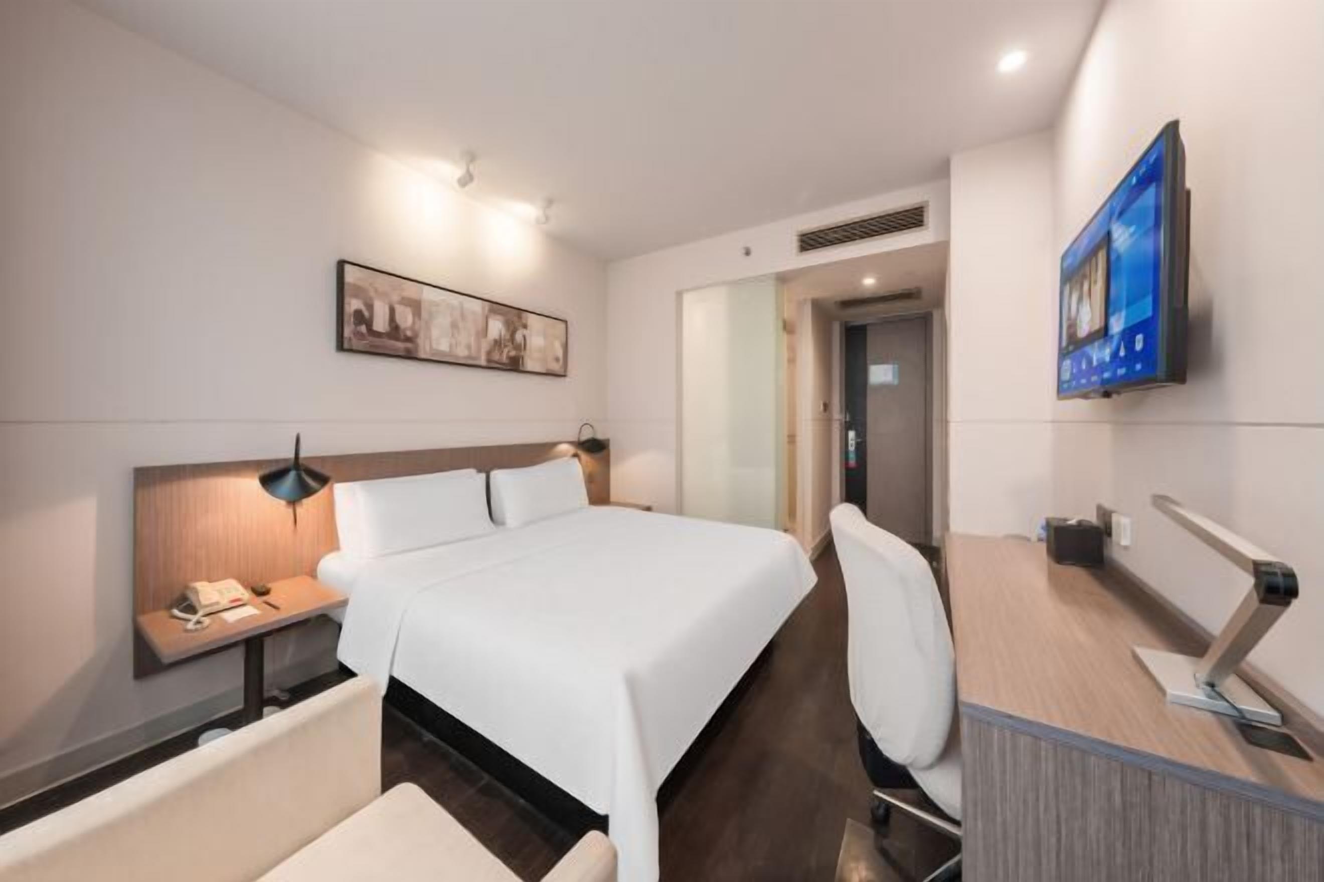 Hotellihuone, jossa on sänky, yöpöytä, tuoli, työpöytä, televisio ja taideteos seinällä.