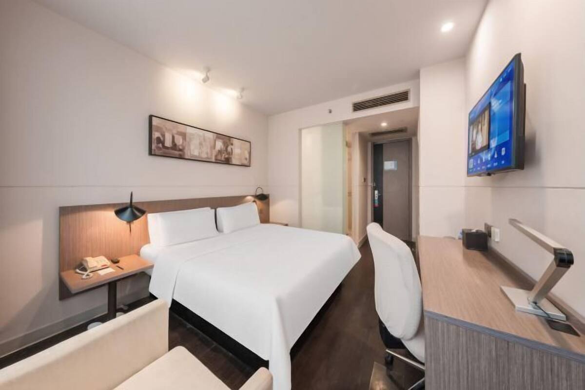 Una habitación de hotel con cama, mesita de noche, silla, escritorio, televisor y una obra de arte en la pared.