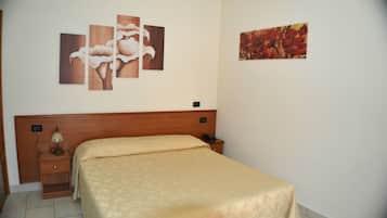 A hotel room with a wooden headboard, a bed with a beige bedspread, a bedside table with a lamp, and two paintings on the wall.