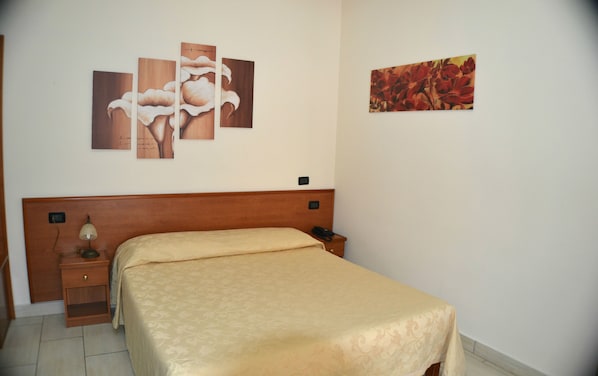 A hotel room with a wooden headboard, a bed with a beige bedspread, a bedside table with a lamp, and two paintings on the wall.