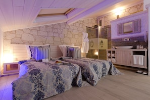 Une chambre avec deux lits, un mur en pierre, une salle de bain avec un lavabo et un grand miroir.