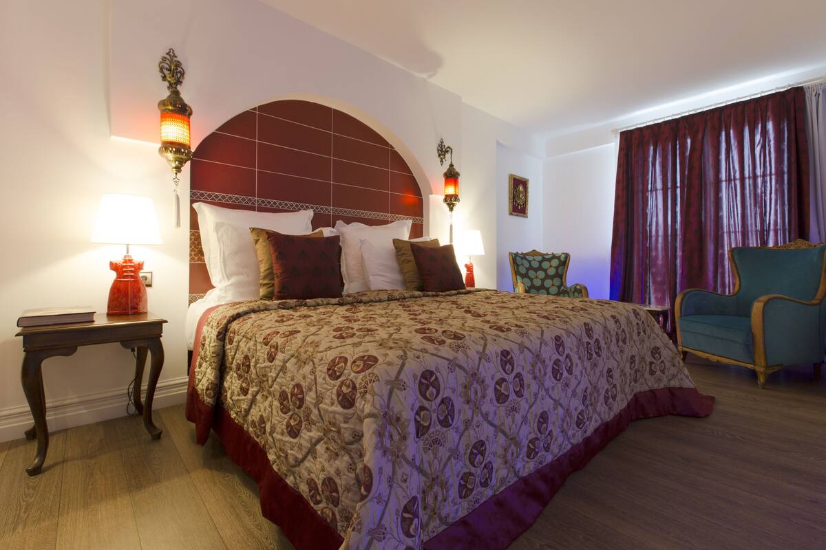 Una habitación de hotel con una cama grande, dos lámparas de noche, una silla y un ventanal con cortinas.