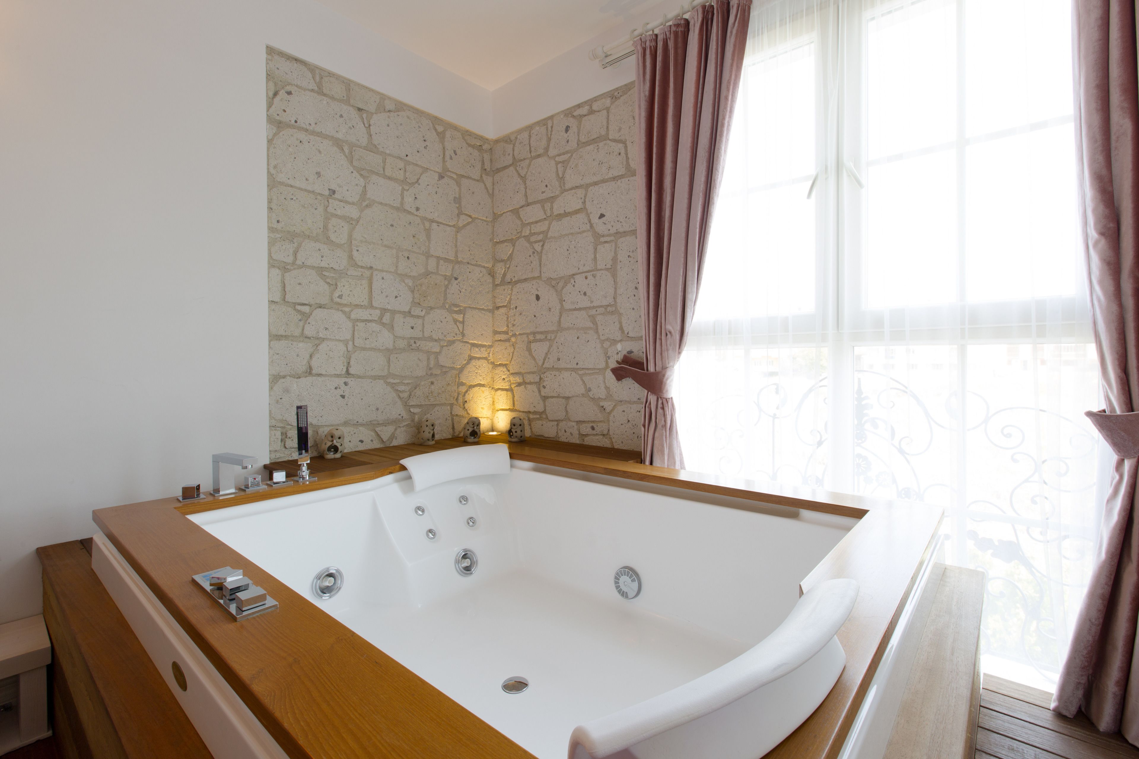 A modern bathroom with a large bathtub, stone wall, and a window with curtains.
