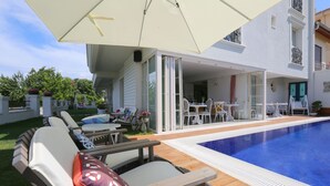 Zona de piscina con sillones de descanso, una mesa y una sombrilla.
