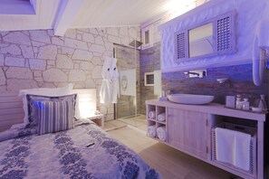 Un dormitorio con una cama grande, un baño con pared de piedra, ducha y tocador con lavabo.