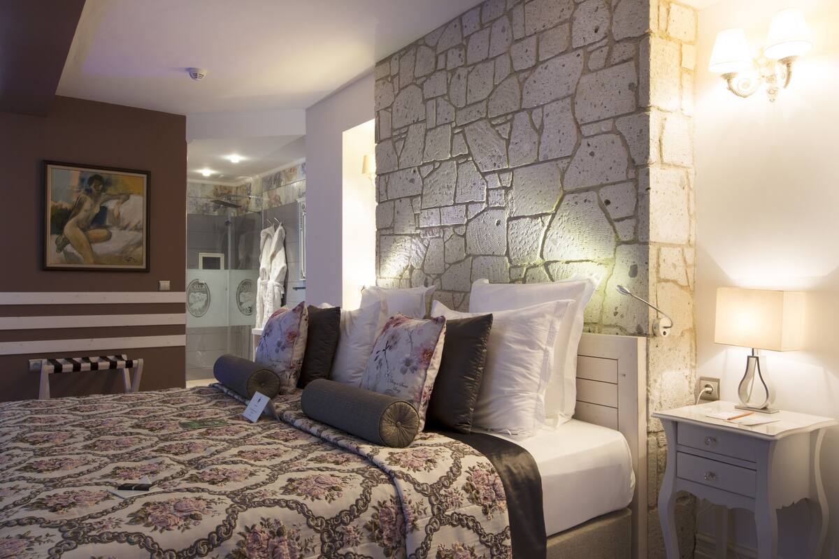 Un dormitorio con pared de piedra, una cama con ropa de cama floral, una mesita de noche con lámpara y un cuadro en la pared.