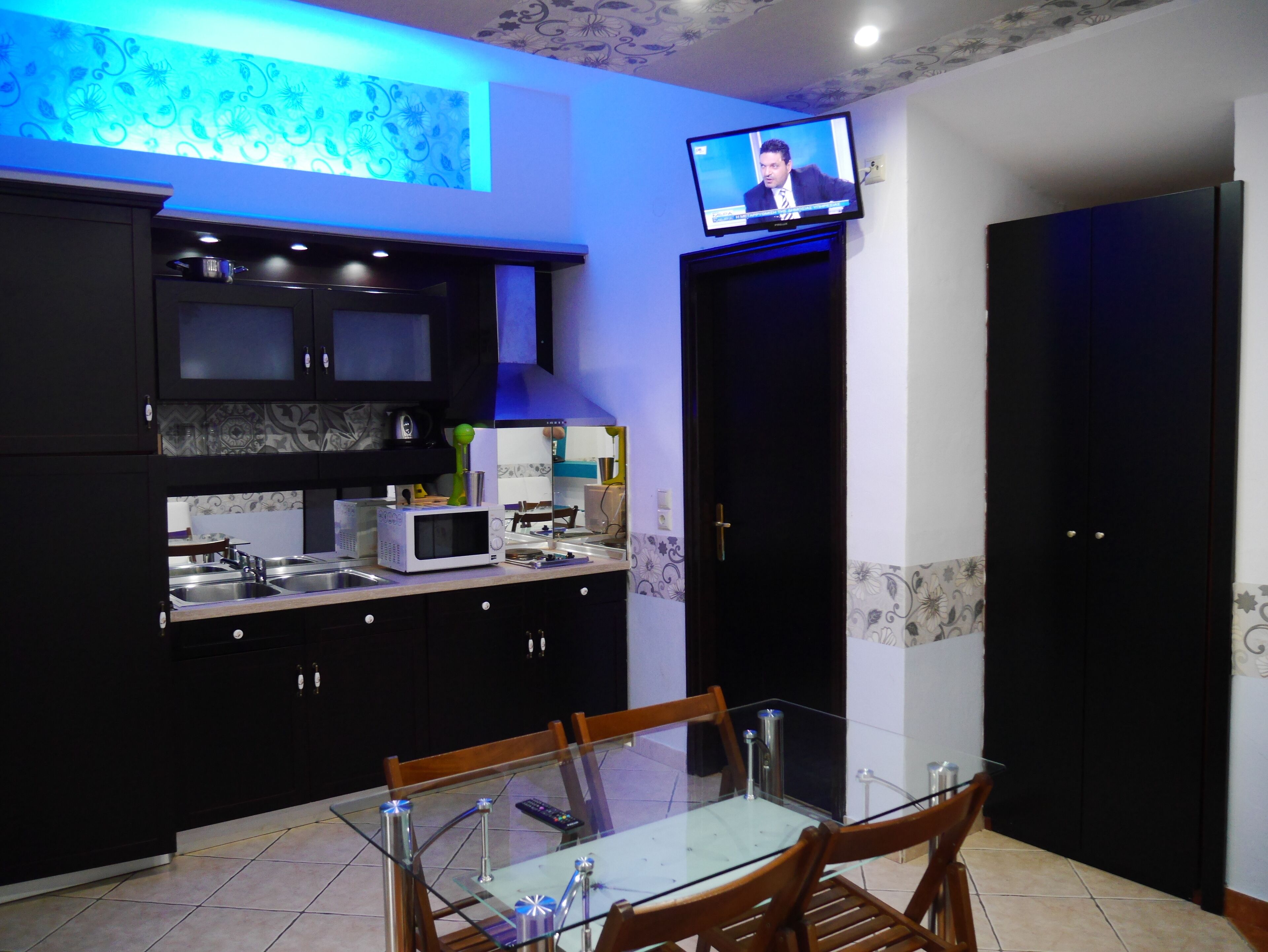 A modern kitchen with dark cabinetry, a glass dining table, and a television mounted above the counter.