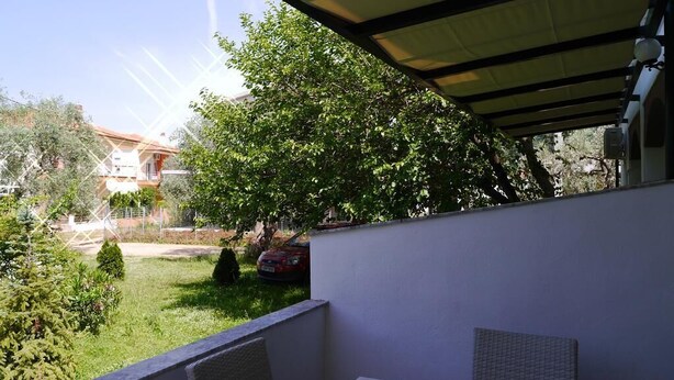 A covered outdoor seating area with a table and chairs, surrounded by greenery and a view of a residential area. - Blue Sky Apartments (Thasos)