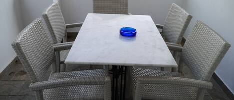 Une table blanche avec un cendrier bleu, entourée de chaises en osier sur un balcon dont le sol est recouvert de pierres.