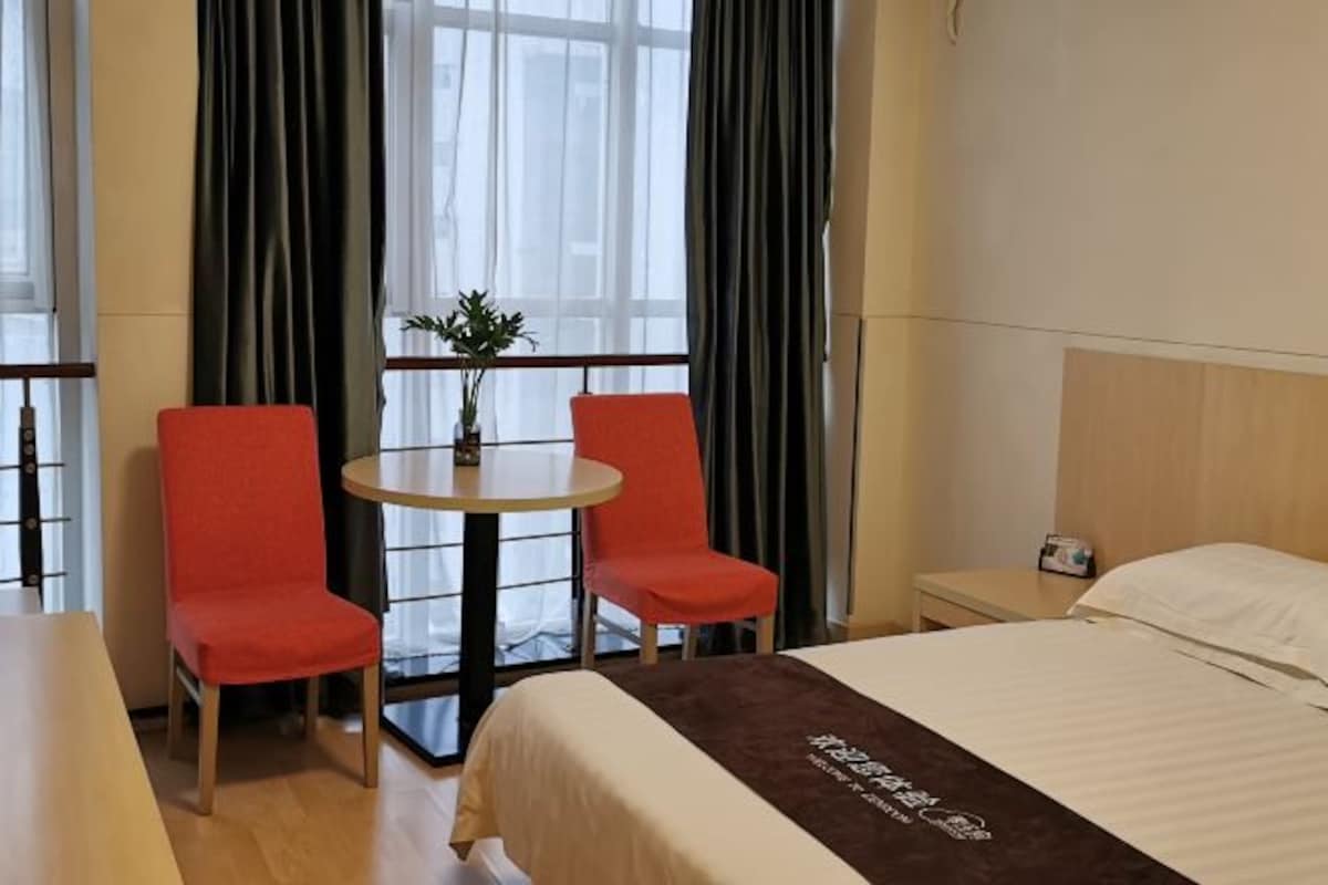 A hotel room with a bed, two red chairs, a small round table with a plant, and a window with curtains.