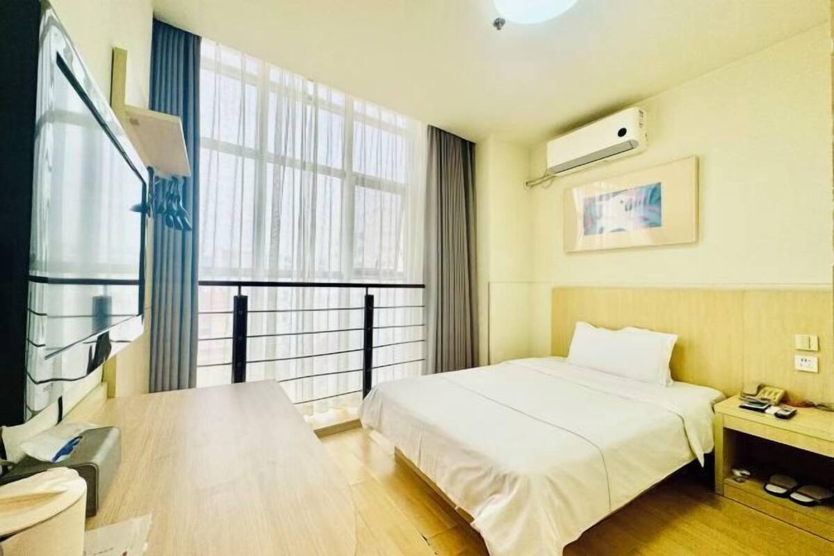 A hotel room with a bed, a desk with a chair, a TV, a window with curtains, a bedside table with a lamp, and a wall-mounted air conditioner.