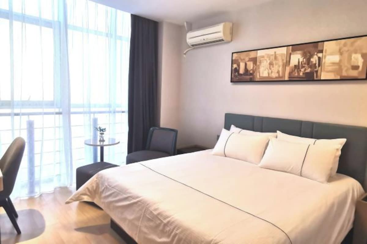 A hotel room with a large bed, a small table, a chair, and a window with curtains.