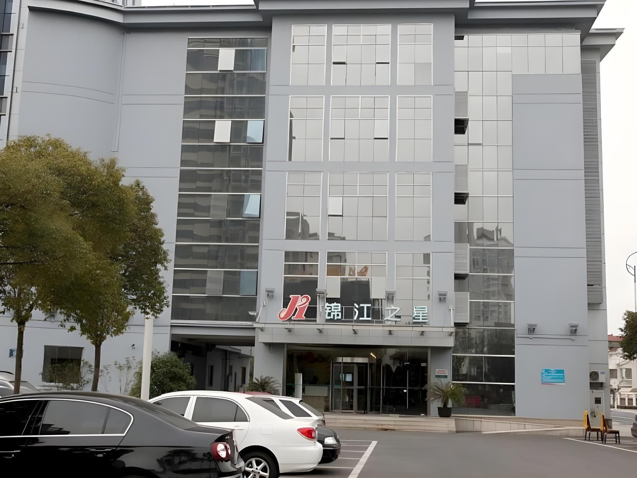 Un edificio moderno con fachada de vidrio, un letrero con caracteres chinos y un estacionamiento con automóviles.