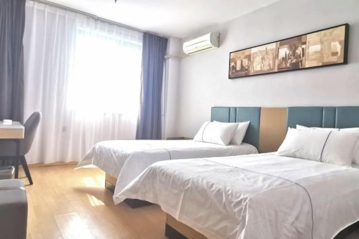 A hotel room with two beds, a desk, and a large window with curtains.