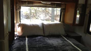 Un lit avec une couverture sombre et deux oreillers, une fenêtre donnant sur des arbres et une tête de lit en bois.
