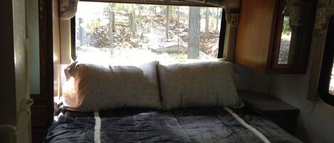 Una cama con un edredón oscuro y dos almohadas, una ventana con vistas a árboles y un cabecero de madera.