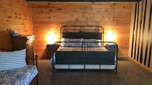 A wooden cabin-style bedroom with a metal bed frame, a bedside table, a dresser, and a small desk with a lamp. - Lake Wanoka Resort (Honesdale)