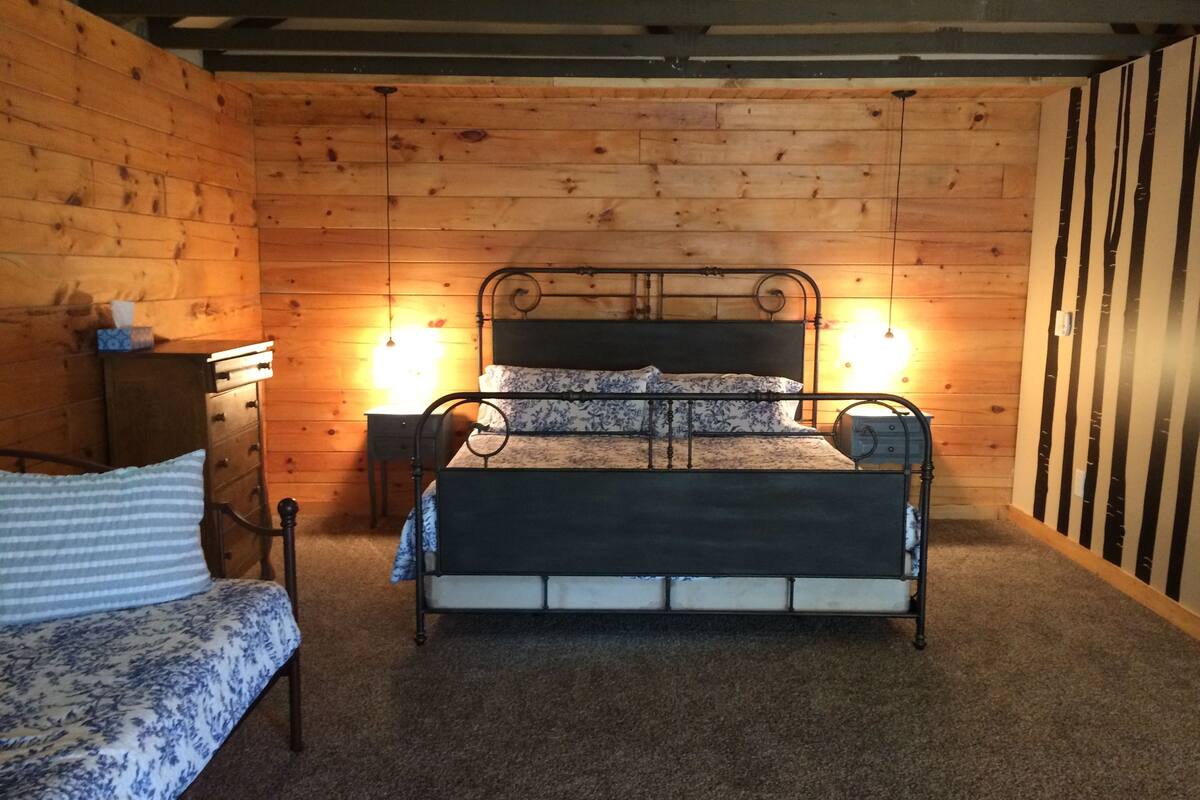 A wooden cabin-style bedroom with a metal bed frame, a bedside table, a dresser, and a small desk with a lamp.