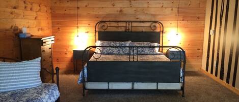 Un dormitorio de estilo cabaña de madera con estructura de cama metálica, una mesita de noche, un aparador y un pequeño escritorio con una lámpara.