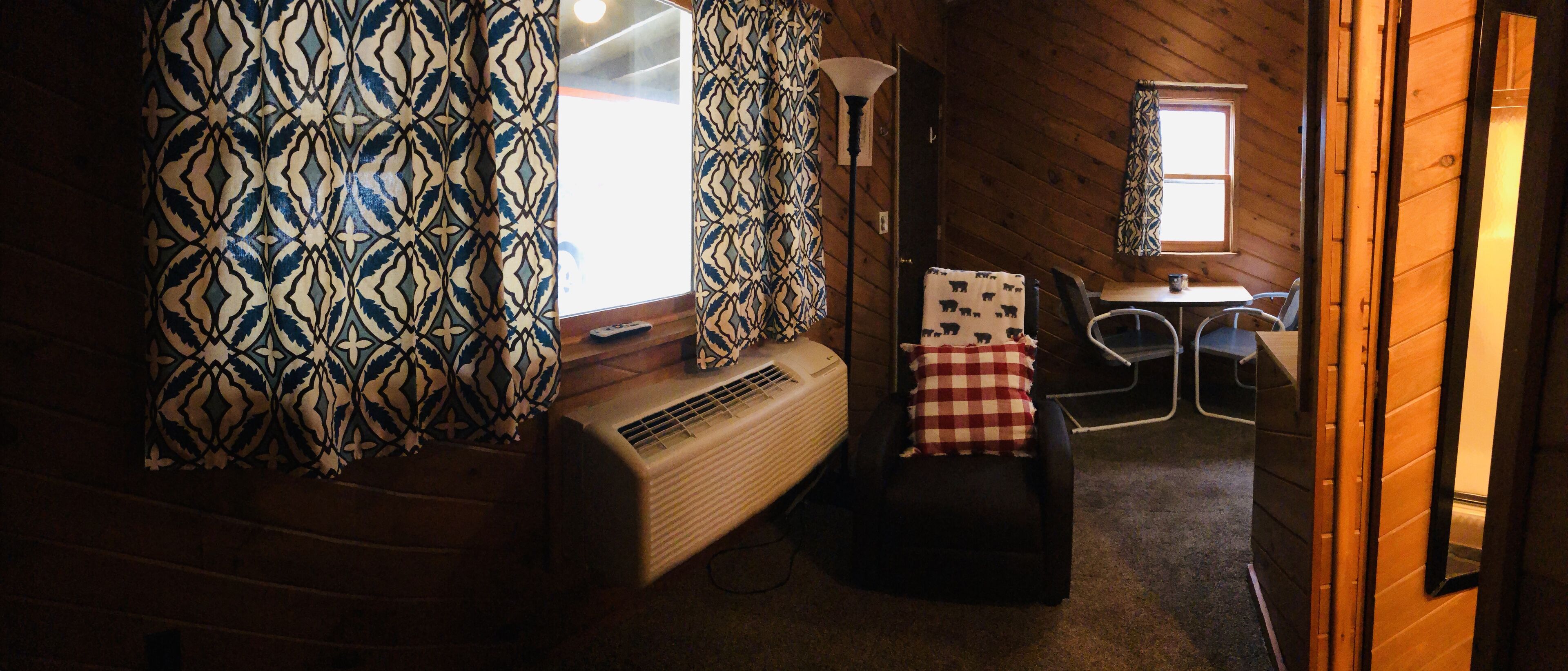 Une pièce avec des rideaux à motifs, un radiateur, une chaise avec un coussin à carreaux et une petite table avec une lampe.