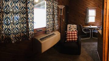 A room with patterned curtains, a radiator, a chair with a checkered cushion, and a small table with a lamp.