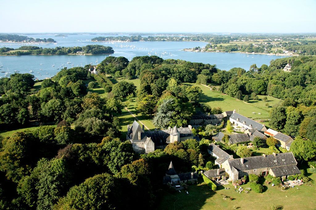 An aerial view of a historic estate with a large body of water, surrounded by lush greenery and a variety of buildings, including a prominent castle-like structure.