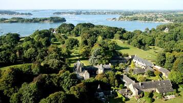 An aerial view of a historic estate with a large body of water, surrounded by lush greenery and a variety of buildings, including a prominent castle-like structure.