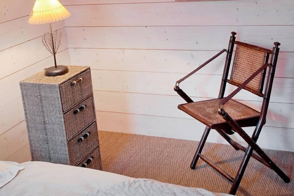 A bedroom with a wooden sloped ceiling, a bed, a dresser with drawers, a chair, and a mirror on the wall.