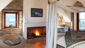 Quarto de loft aconchegante com vigas de madeira aparentes, lareira, cama e poltrona de vime.