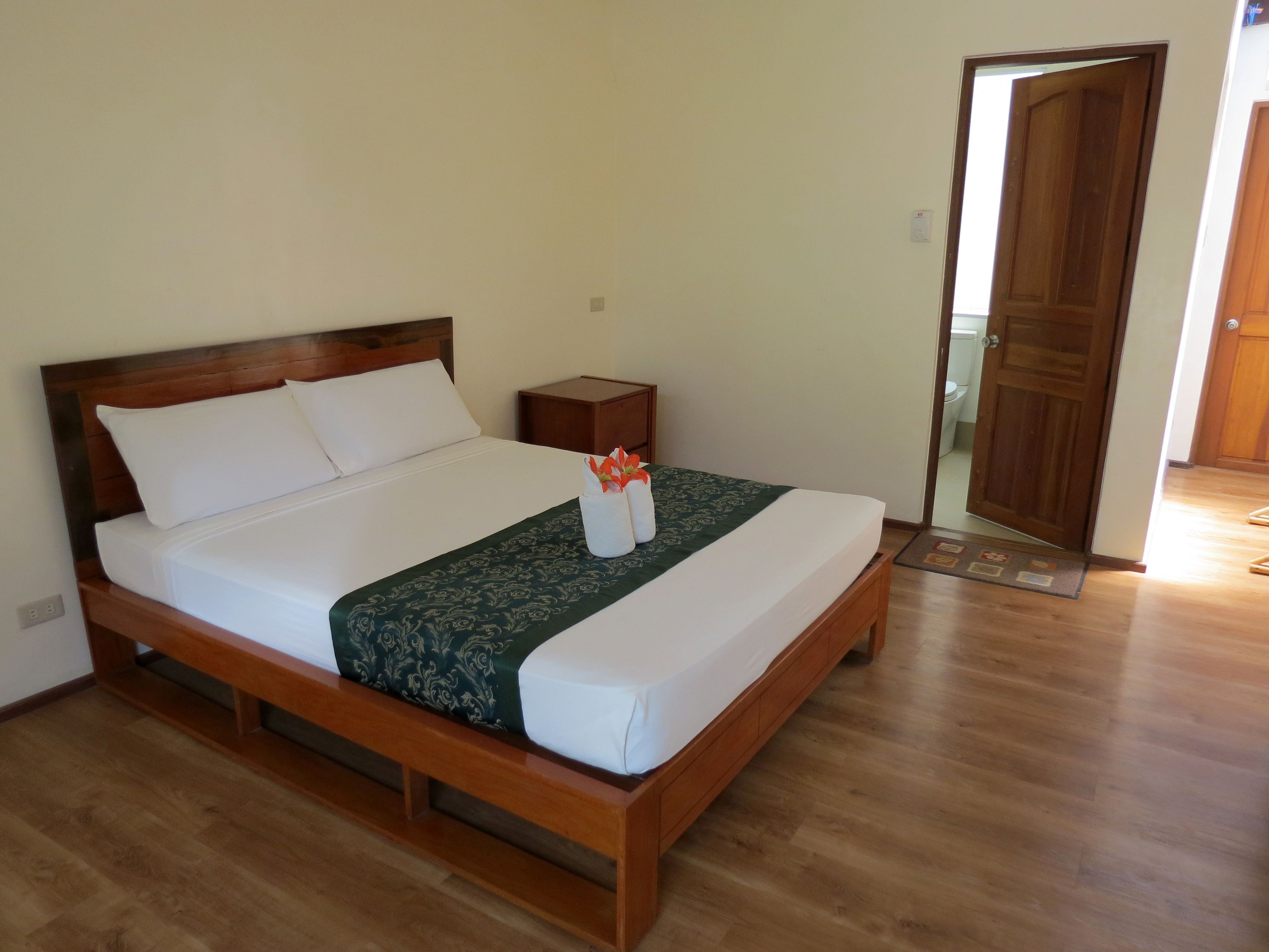 A neatly made bed with white linens and a green patterned bedspread, a wooden nightstand with a small lamp, and a wooden door in the background.