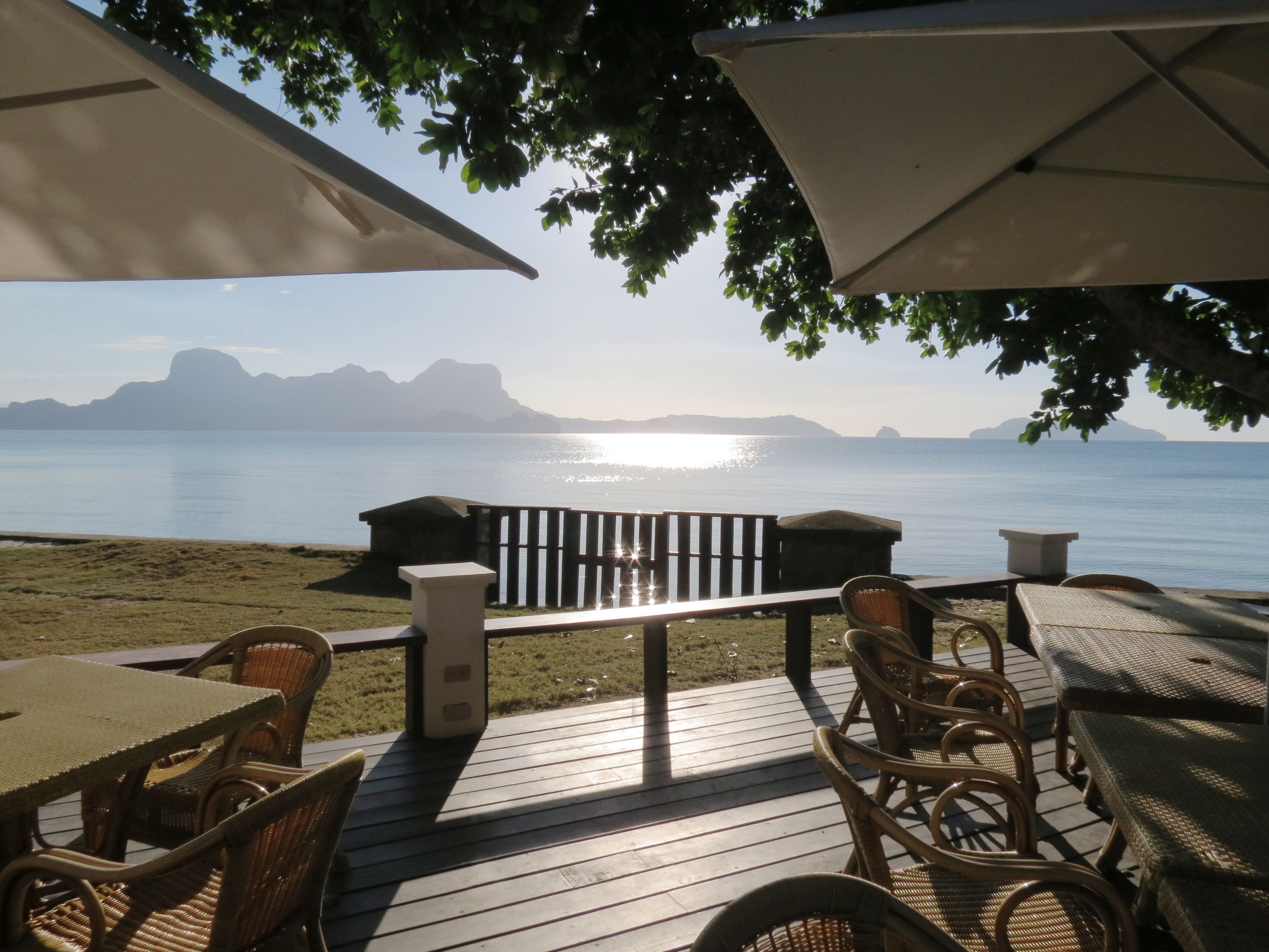 A deck with wicker furniture and umbrellas overlooking a calm sea with distant mountains.