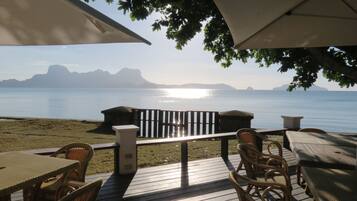A deck with wicker furniture and umbrellas overlooking a calm sea with distant mountains.