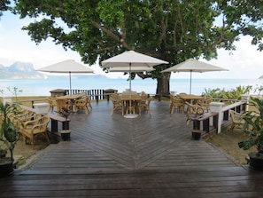 An outdoor dining area with wooden tables and chairs, surrounded by umbrellas, overlooking a body of water and mountains.