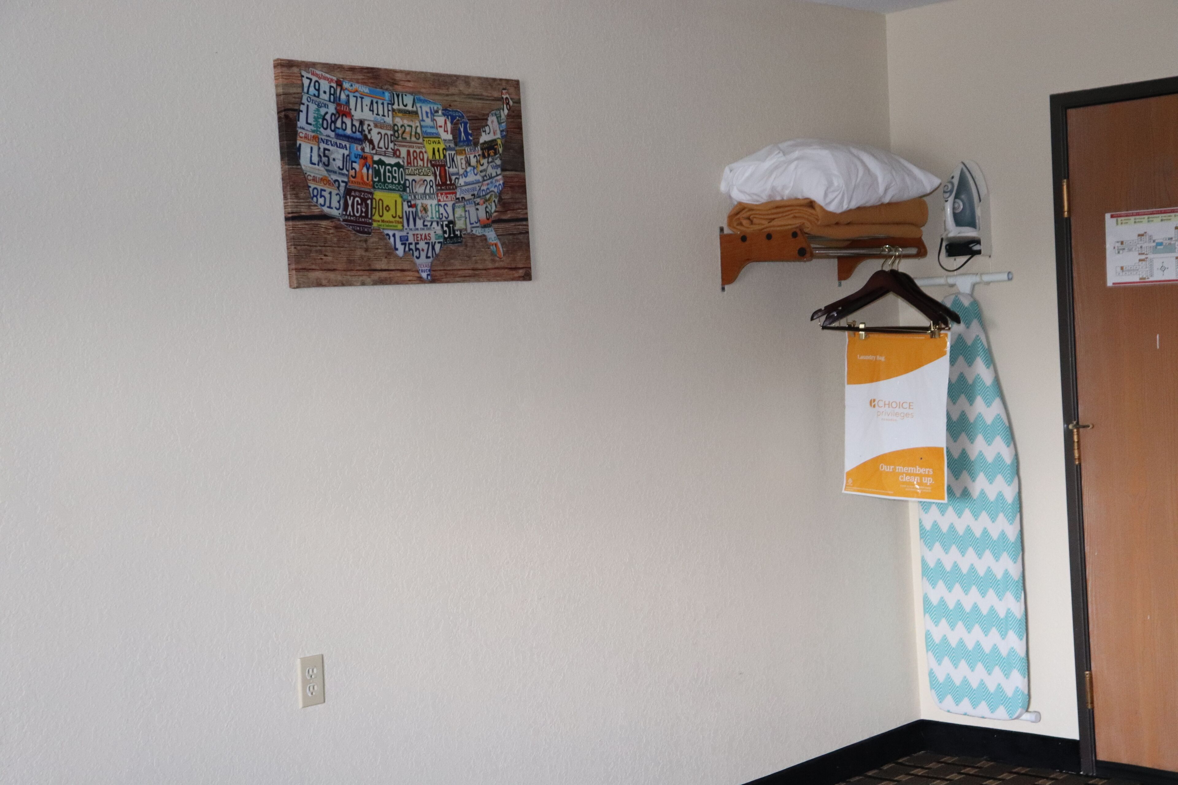 A room with a wooden shelf holding a pillow and an iron, a wall-mounted ironing board with a tie, and a map of the U.S. on the wall.