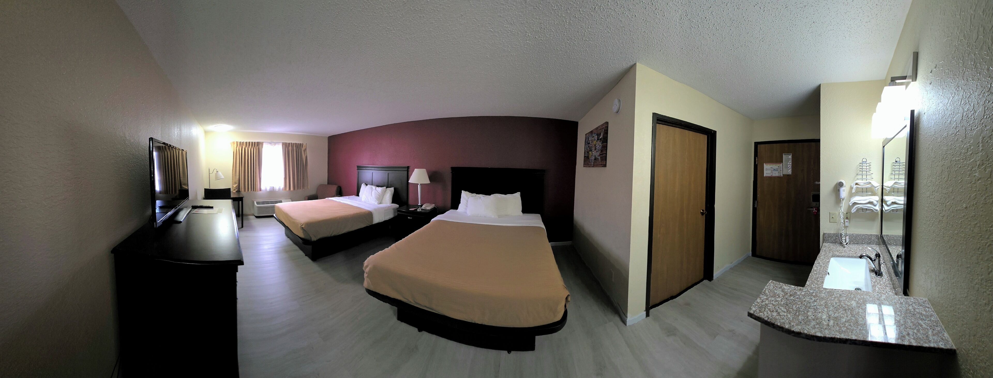 A hotel room with two beds, a desk, a TV, a mirror, and a bathroom with a sink and mirror.