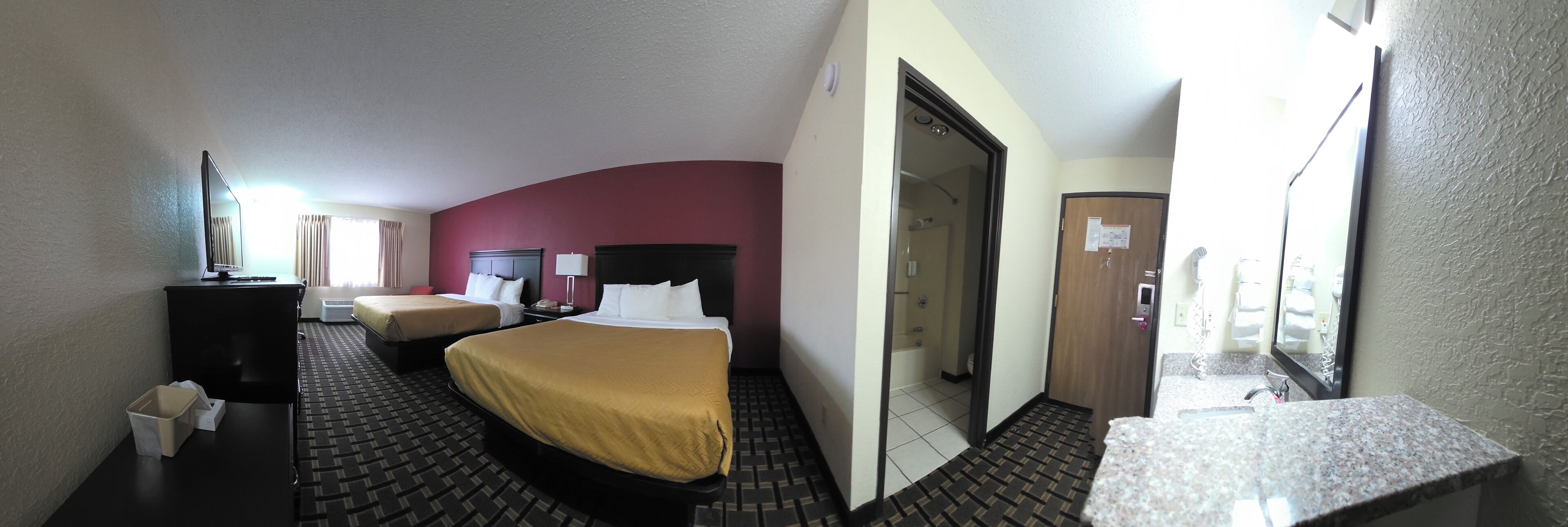 A hotel room with two beds, a desk, a chair, a small table, a mirror, and a bathroom visible through an open door.