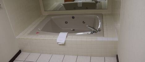A bathroom with a large bathtub, a mirror above it, and a tiled floor.