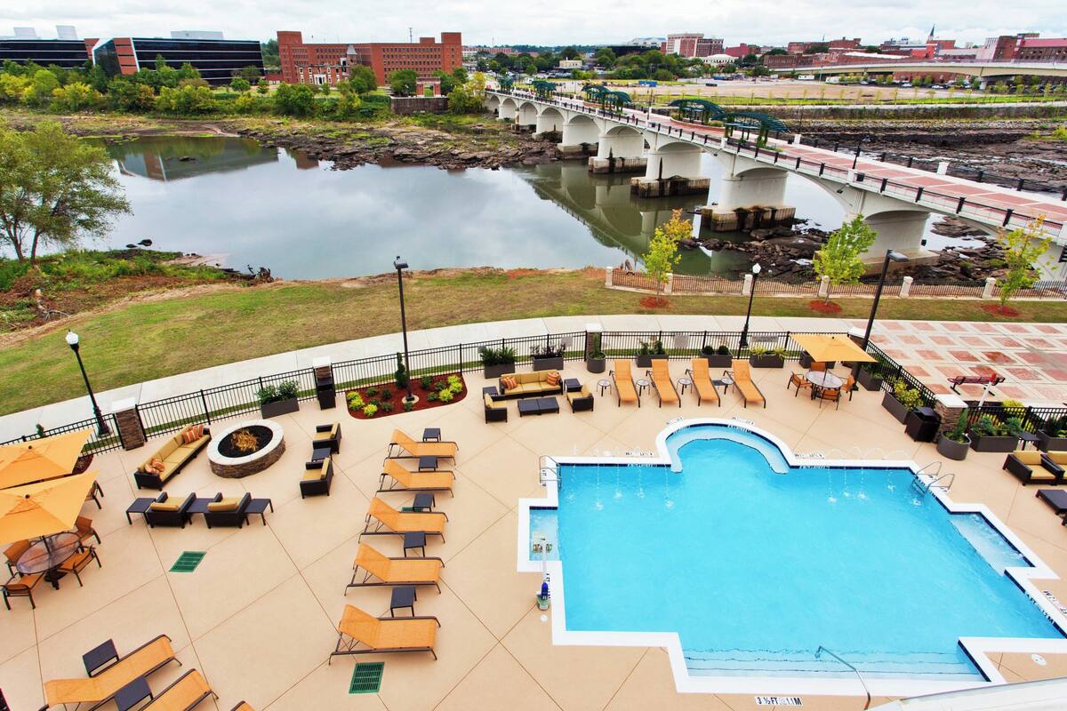 A rooftop pool area with lounge chairs, a fire pit, and a view of a bridge over a river.