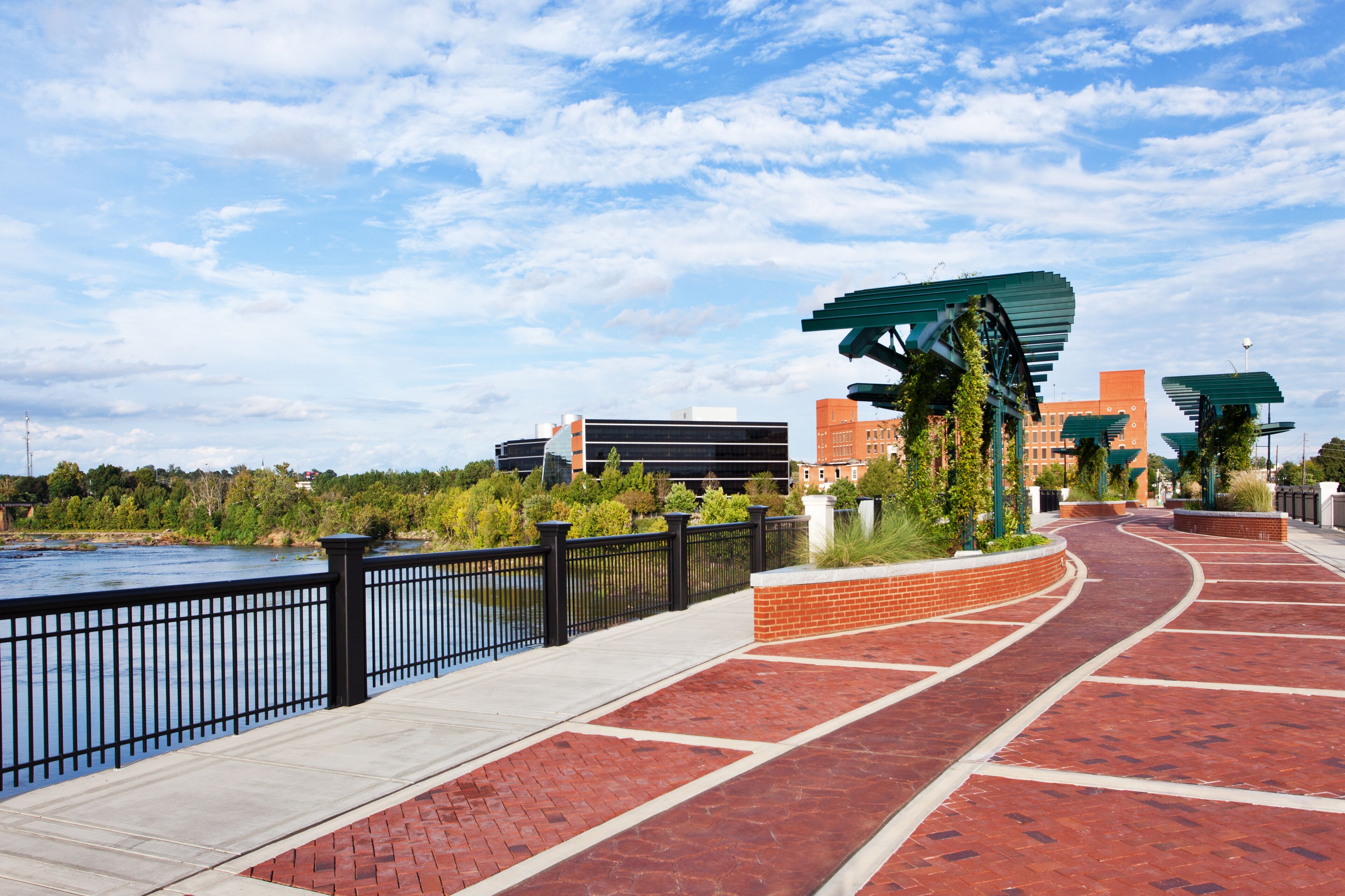 A waterfront walkway with a brick path, metal railing, and green canopies.