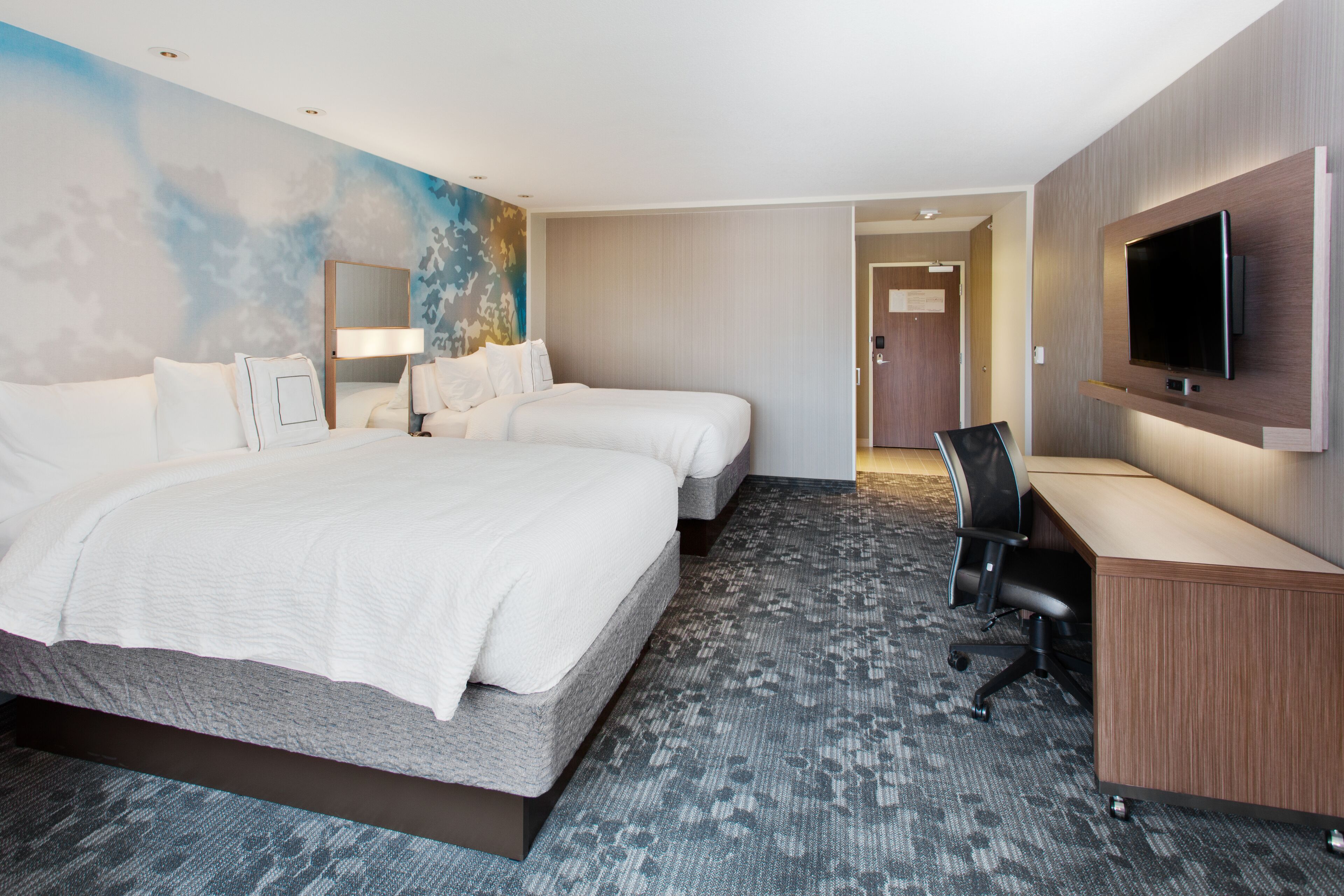 Habitación de hotel con dos camas, un escritorio, televisor y piso alfombrado.